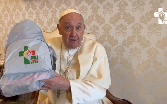 Pope Francis holds up a gray backpack with the World Youth Day logo as he speaks in a room with the Vatican City emblem on the cream wallpaper