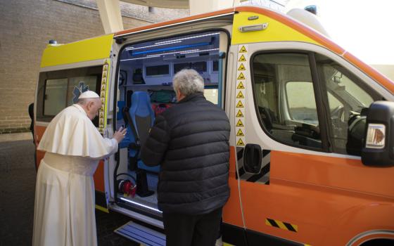 Pope Francis raises his hand as he stands outside an orange and yellow vehicle next to a man with white hair