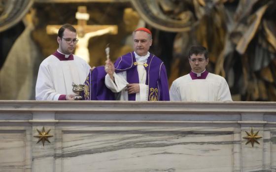 A white man wearing a red zucchetto uses an aspergillum to sprinkle holy water on an alter. Two men in clerical collars stand beside him.