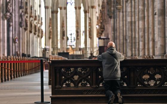 A man kneels on a kneeler in an empty-looking church with larger pillars lining the sides