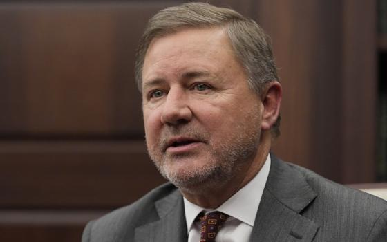 A white man with gray stubble wearing a suit and tie looks into the camera