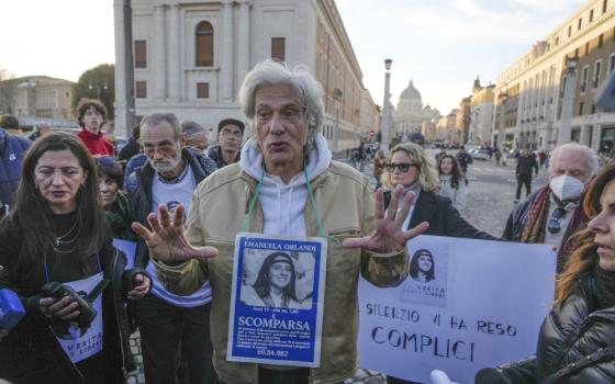 An older white man, surrounded by a crowd, talks with his hands and wears a sign around his neck with a picture of a girl on it