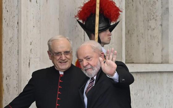 An older white man in a suit waves at the camera next to a man wearing a cardinal's cassock with a member of the Swiss Guard in the background