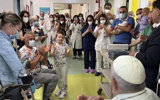 A large group of people, adults and children, many wearing masks or holding cellphones, stand around Pope Francis
