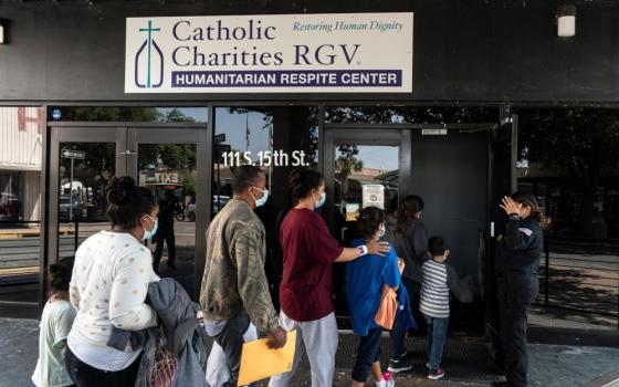 A line of people wearing masks walk into a building with the sign "Catholic Charities RGV Humanitarian Respite Center"