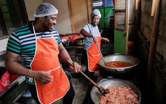 Two Black people wearing hair nets and orange aprons smile and stir food in large pots