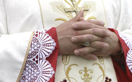 A white man's hands are folded as he stands in intricately embroidered white vestments. His head is not visible.