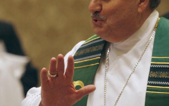 A white man with a moustache wears a violet zucchetto and green stole over white vestments as he talks and gestures