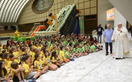 Pope Francis waves and uses a cane to walk across the front of a large group of children wearing t-shirts seated on the floor. An inflatable Titanic and other inflatable slides are visible in the background