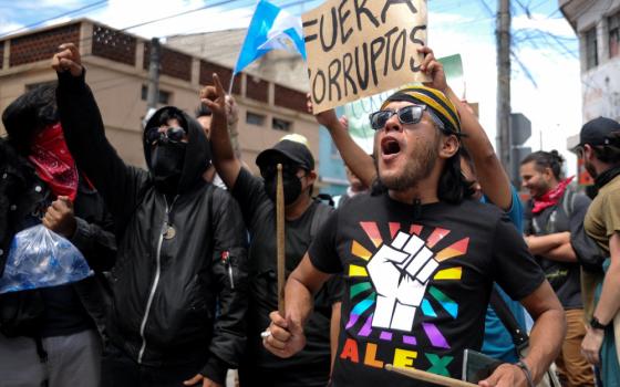 A group of people march in the street. Some wear masks. One holds a sign saying "Fuera Corruptos" high.