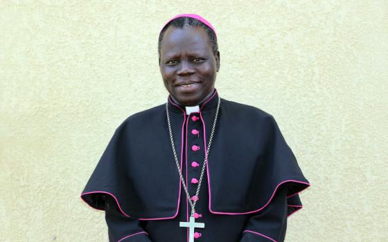 A Black man stands before a white wall and wears a violet zucchetto, pectoral cross and bishop's cape