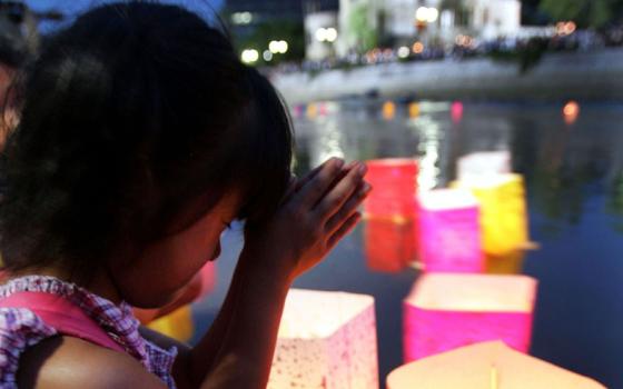 A Japanese child squats and presses her palms together and thumbs to her forehead in front of a waterway with floating paper lanterns. A white building is blurry in the background