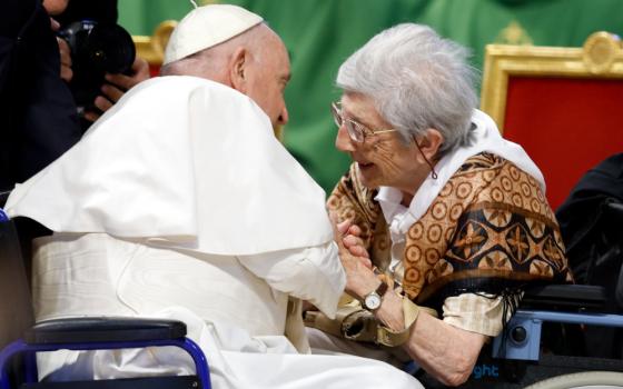 Pope Francis, seated in a wheelchair, leans forward to clasp the hands of an older white woman sitting in a wheelchair 