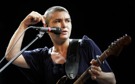 A white woman with a buzz cut holds a guitar and sings into a microphone while making a hand gesture pointing down