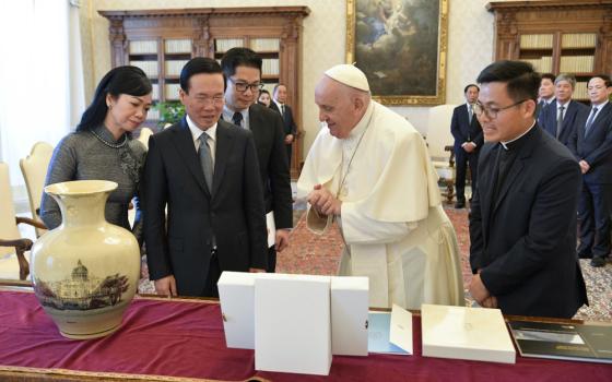 Pope Francis stands between a Vietnamese man in a suit and an Asian man in a clerical collar as they study objects on a table