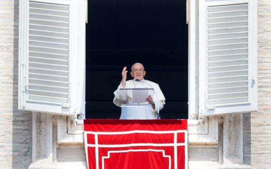 Pope Francis stands at his apartment window and raises one hand as he speaks behind a clear lecturn