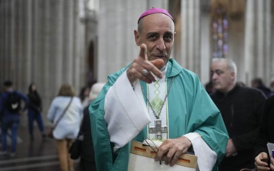 A man wearing green vestments and a violet zucchetto points at the camera while standing in a church