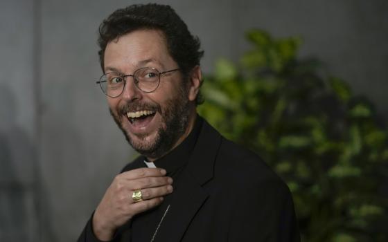 A younger man wearing glasses and a clerical collar beams at the camera while touching his collar