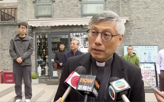 An East Asian man wearing a clerical collar and glasses speaks into different TV microphones