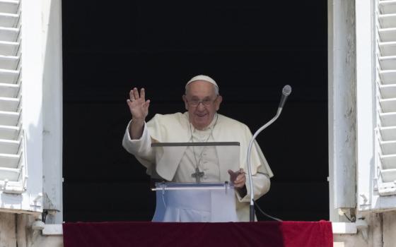 Pope Francis lifts his hand as he stands in his apartment window behind a clear lectern and microphone