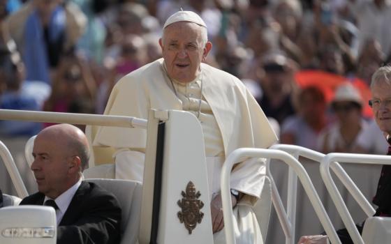 Pope Francis stands in the back of the popemobile. A crowd is visible and blurred behind him.