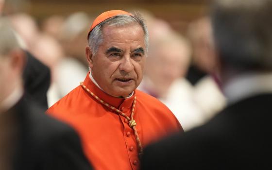 An older man wearing a red zucchetto and a red choir dress vestment stands amid blurred out people