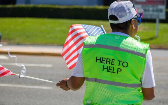 The back of a person on a street, holding American flags and wearing a green vest that says, "HERE TO HELP." (Unsplash/Elissa Garcia)