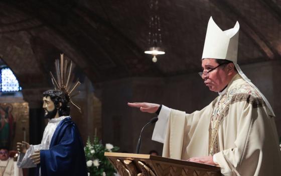 A man wearing glasses and white mitre and vestments stands at a lectern and speaks with an outstretched hand