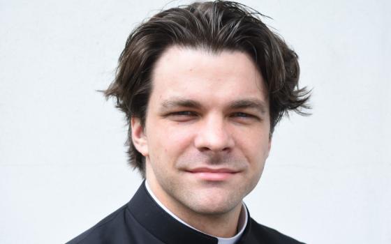 A white man wearing a clerical collar with styled, swoopy hair looks into the camera