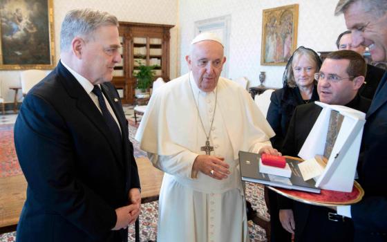An older white man in a suit stands next to Pope Francis, who places his hand on an artwork another man is holding towards the first man
