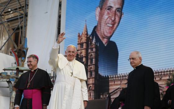 Pope Francis raises his hand to wave while standing in front of a large image of a white priest