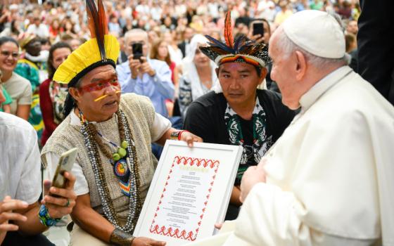 Two men wearing feather headpieces, one with a nose piercing, hand Pope Francis a frame with language