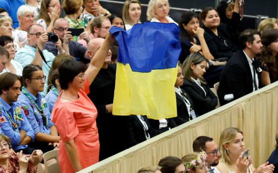 People in a sitting crowd stand with a Ukrainian flag, including a Brown woman with an orange dress