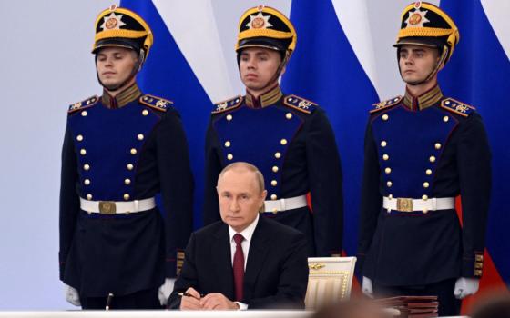Vladimir Putin, in a suit, sits in front of three Russian soldiers wearing dress uniforms