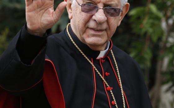 An older white man wearing cardinal's clothes and glasses transitioned to sunglasses raises his hand to wave.