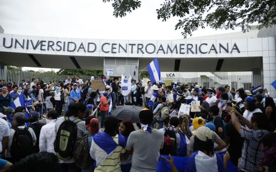 A group of people carrying Nicaraguan flags stand in front of an arch that reads "Universidad Centroamericana"