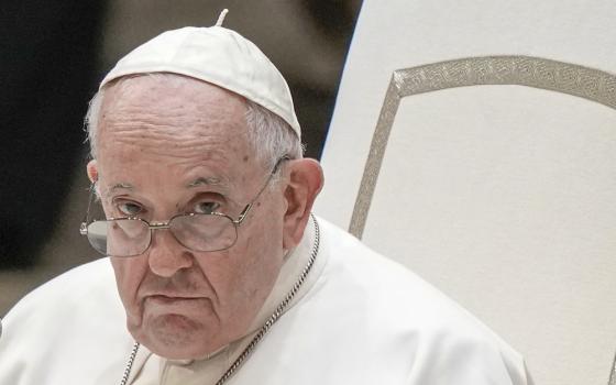 Pope Francis' head and shoulders are seen against a white chair. He wears glasses.