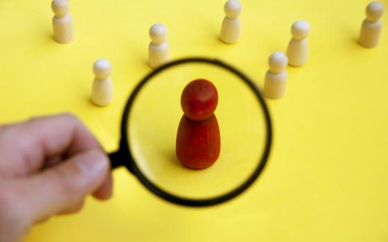 A hand with a magnifying glass singles out a red-painted wooden figurine among seven unpainted figurines (Dreamstime/Mauriceyom98)