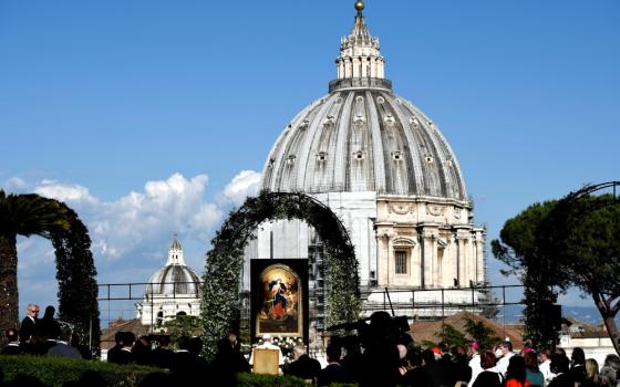 Leafy green arches are visible in front of the dome of St. Peter's Basilica with people sitting in the garden in front of an image of Mary