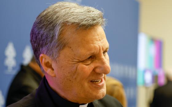 A gray-haired white man wearing a clerical collar smiles. The Vatican press conference backdrop is blurry in the background.