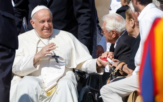 Pope Francis waves his hand while sitting in his wheelchair. With his other hand, he holds the hand of an older man in the crowd to the side.