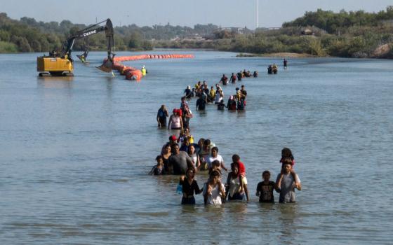 People, many with children on their shoulders, walk out into the middle of a river. A construction machine and buoys can be seen behind them.