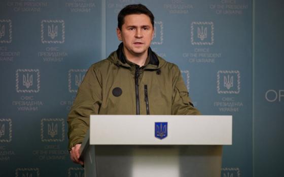 A white man with short dark hair wearing a green jacket speaks at a podium