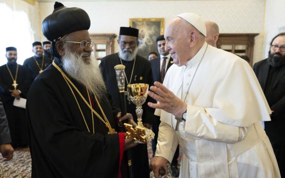 Pope Francis smiles at an older brown man with a beard wearing a black hat in a Hershey kiss shape, black robes, and long necklaces.