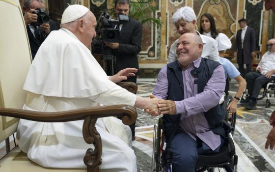 Pope Francis, sitting in a chair, shakes the hand of a man using a wheelchair