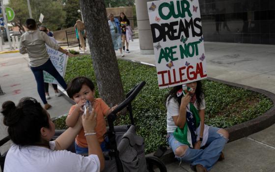 A person sitting cross-legged holds a sign that says "Our dreams are not illegal," while next to a person feeding a toddler.