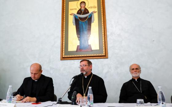 Three men wearing clerical collars sit at a table below a painting of a woman saint
