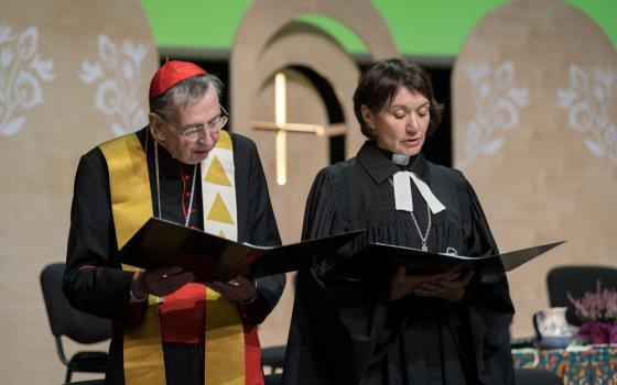 A man wearing a red zucchetto and a yellow stole stands next to a woman wearing a clerical collar and a black robe