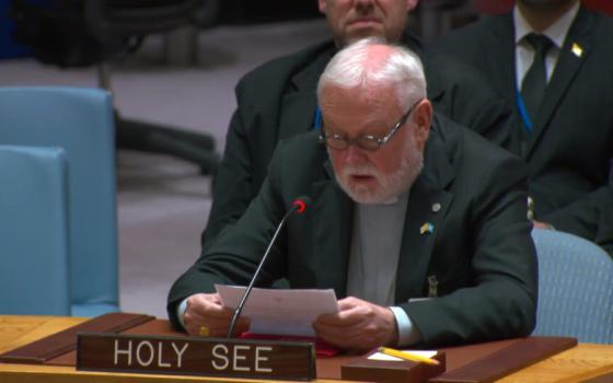 A white-haired white man with a beard and glasses wearing a gray shirt and clerical collar and black suitspeaks into a microphone behind a plaque saying "Holy See"