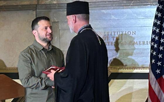 President Volodymyr Zelenskyy, wearing green, hands a red box to a white man wearing a cylindrical black hat and black vestments